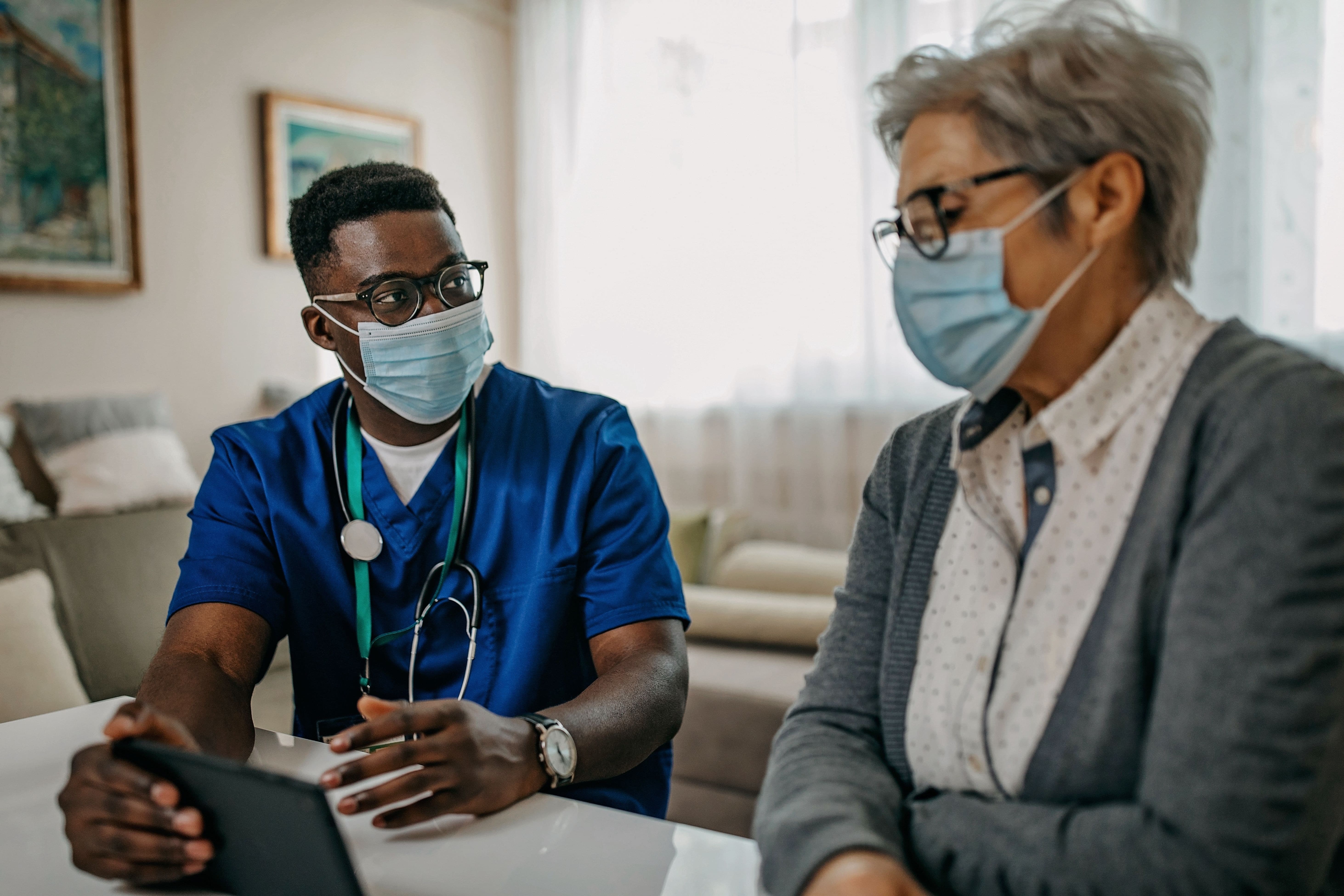 Nurse showing patient a screen in her home with masks on. Nurse showing patient a screen in her home with masks on.